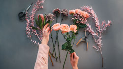 A woman's hands and arms over various floral and leafy stems with floral arranging supplies.